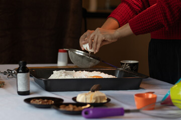 Unrecognizable dark-skinned woman breaking an egg and pouring it into a tray with flour, along with ingredients and kitchen tools, in preparation of a baking recipe.