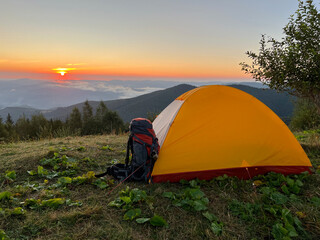 Tourist adventure backpack, packed hiking travel backpack stands near the tent on high hill against backdrop of mountains. Filled tourist hiking backpack that stands on the grass. Sunrise in mountains