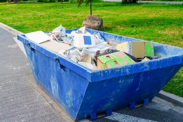 A blue dumpster is sitting in the park. The dumpster is filled with various types of garbage...