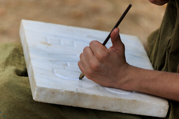 Stonemason using a chisel to carve roman letters into a white marble block that he is resting on his knees. Close up of a mans hand using a chisel. 