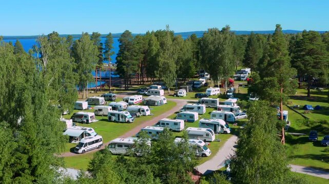 Campsite or campgrounds in a green forest and blue lake behind. camper van mobile home driving. Sunny summer day filmed in aerial view in Sweden Scandinavia