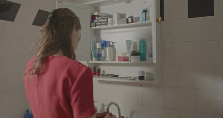 Woman in a red dress packing toiletries from medicine cabinet in white bathroom