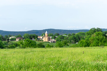 Field with green grass, edge of forest, brick church of Kodalo village. Mountains and clear blue sky in the background