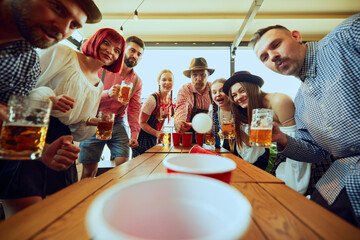 Group of people cheerfully playing beer pong, having fun and bar while celebrating beer festival. Bavarian traditions. Concept of Oktoberfest, festival, party, brewery, traditions