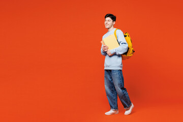 Full body side view young smiling happy man student he wear blue casual clothes backpack bag hold books walking going isolated on plain red orange background. High school university college concept.