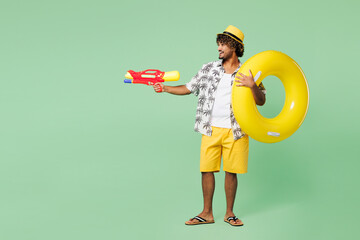 Full body side view young Indian man wear beach shirt hat hold rubber ring rest near hotel pool play shoot from water gun isolated on plain green color background. Summer vacation sea sun tan concept.