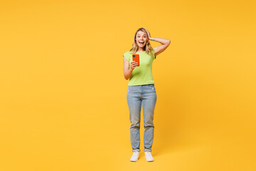 Full body young happy shocked surprised blonde woman wear green t-shirt casual clothes put arm on head hold in hand use mobile cell phone isolated on plain yellow orange background. Lifestyle concept.