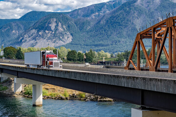 Red classic American bonnet big rig semi truck transporting cargo in refrigerator semi trailer running on the transportation bridge beside rail road bridge across the river in Columbia Gorge area