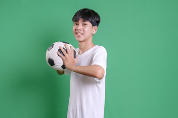 Young Asian Man Wearing White T-shirt Holding Soccer Ball And Looking At Camera Isolated On Green...