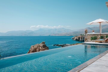 Fototapeta premium Elegant infinity pool overlooking the sea in Kalythes, Crete, with clear blue skies, distant mountains, and luxurious villas in the background, featuring a stone wall and a white umbrella.