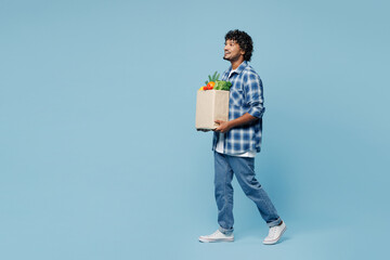 Full body side profile view fun young Indian man wear shirt hold craft bag for takeaway mock up with food products walk go isolated on plain blue background. Delivery service from shop or restaurant.