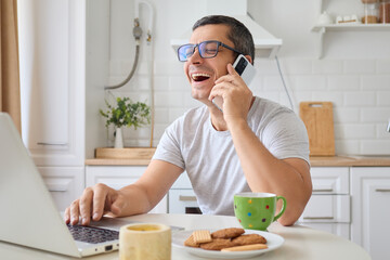 Laughing man freelancer working alone at home using laptop and smartphone for online work and phone calls happy with funny communication in kitchen managing remote projects and social media