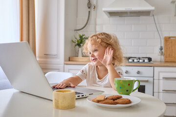 Funny curly haired little girl having video call on computer at home enjoying internet communicating waving hand hello gesture sitting at table in kitchen