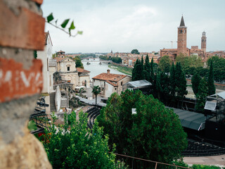 A scenic view of a historic Italian town along the river featuring towers, greenery, and an outdoor stage during a cloudy afternoon