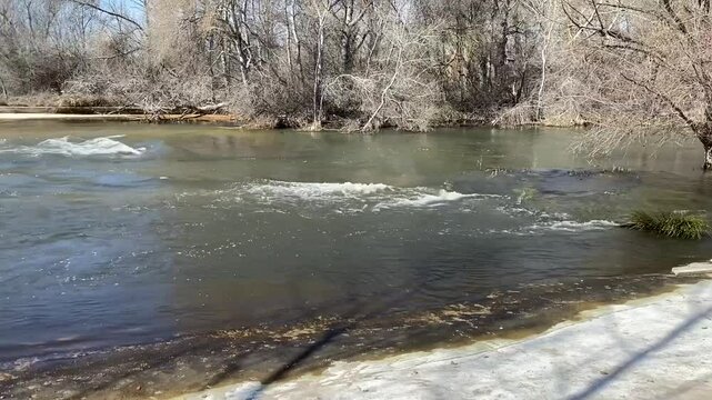 Recorrido visual en la orilla de un rio con caudal de agua y piedras, al que le atraviesa un puente met&aacute;lico naranja, paso peatonal, senderos o v&iacute;as pecuarias, paisaje natural, solitario, d&iacute;a soleado