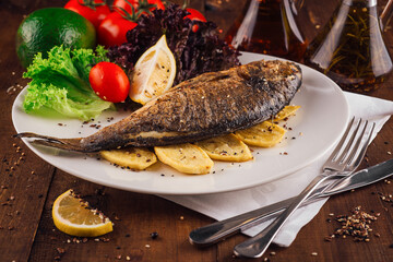 Grilled fish with potatoes and vegetables on white plate being served on wooden table