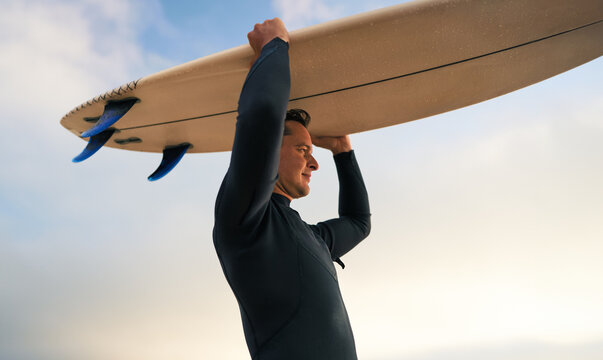 A surfer in a wetsuit holding a surfboard above his head at the beach during sunset. Ready to catch waves.