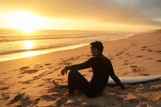 A surfer in wetsuit sits on the sandy beach with a surfboard at golden sunset, reflecting on the serene ocean view.