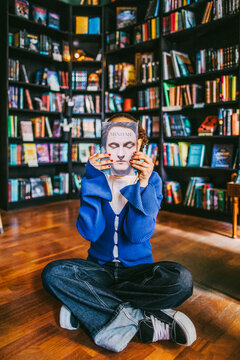 Young woman hiding face with book while sitting cross-legged in bookstore