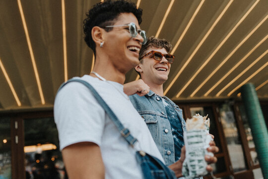 Low angle view of happy young man in sunglasses leaning on male friend holding wrap sandwich outside shop