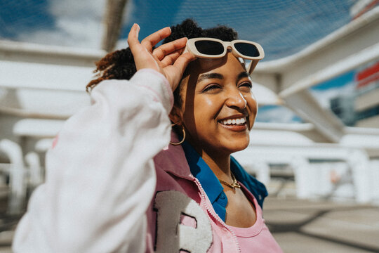 Happy fashionable curly hair woman in letterman jacket holding sunglasses at parking lot
