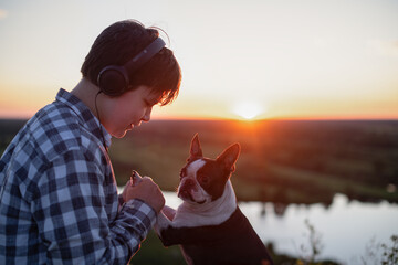 A teenage boy on a walk in the fresh air with his dog and wearing headphones is relaxing alone, enjoying the summer sunset views. Conscious time for mental and emotional health.