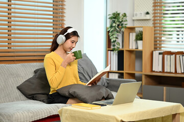 Fototapeta premium Young asian woman in headphone drinking hot tea and reading book on sofa at home