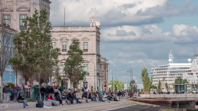 Seaside street Avenida Ribeira das Naus next to Praca do Comercio timelapse in Lisbon, Portugal. People sitting and walking on the waterfront near Commercial square. Cloudy sky and port on background