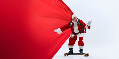 Santa Claus riding skateboard while holding large red cape, with enthusiastic expression against white studio background. Concept of winter holidays, Holly Jolly mood, celebration. Ad © Lustre Art Group 