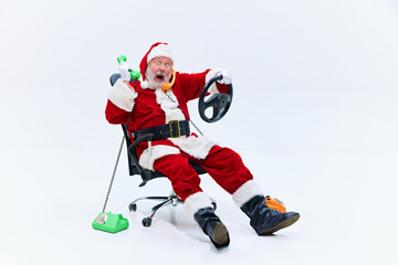 Santa chats on two phones, green and orange, while gripping steering wheel, his expression mix of chaos and frustration, reclining in his office chair. Concept of winter holidays, celebration. Ad