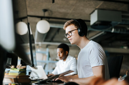 Young male programmer doing coding on computer with wireless headphones in office