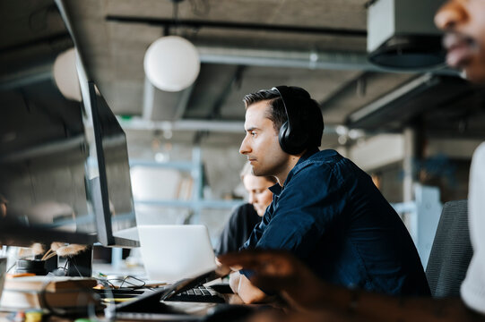 Young programmer with wireless headphones sitting and looking at computer in office