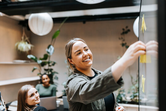 Smiling female business professional sticking adhesive note on whiteboard at startup office