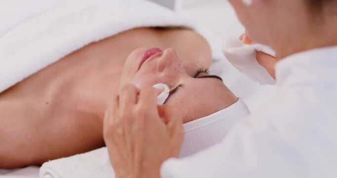 Close-up photo pf professional cosmetologist or beautician doctor hands making anti-age procedures cleaning with napkins facial mask of young pretty woman client in beauty spa clinic. 4k video.