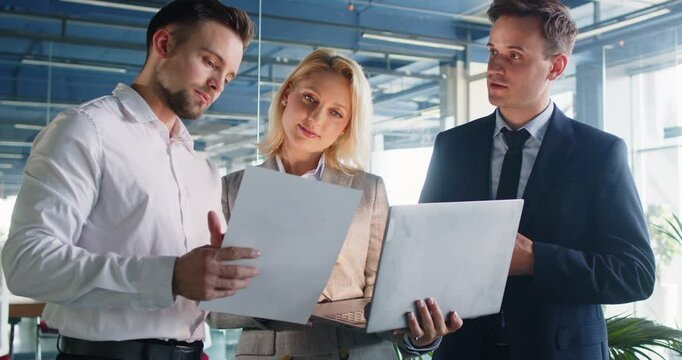 Young business team in the office workplace. Group of people having a work discussion. Three colleagues, one woman and two men, standing in the office, discussing a financial contract, using a laptop