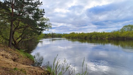 In early spring the river overflows, the water is high and muddy. A fast current floods the floodplain and bushes on the banks. The water reflects the trees and bushes with young leaves and the sky