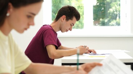 Obraz premium High school teenagers in classroom, girl sit at desk writing test, looks at camera smiles and shows thumb up. Modern education concept.