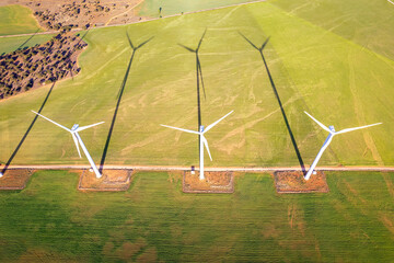 Directly above view of of three towering wind turbines casting long shadows over lush green fields,...