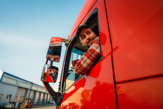 A truck driver leans out of the window of a red truck while parking on a parking lot in an industrial area with buildings in the background.