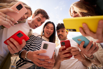 Diverse group young people in circle staring in surprise at cell phone screen together outdoors. Amazed generation z student friends using colorful mobiles enjoying interesting social media content 