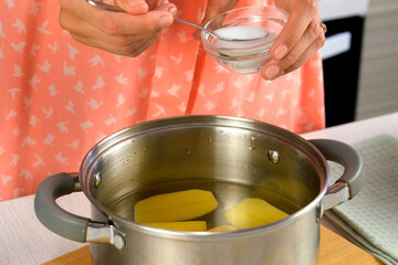 Woman adding salt to water with peeled potatoes cut on halves in saucepan to cook, boil on kitchen...