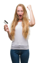 Naklejka premium Blonde teenager woman holding credit card annoyed and frustrated shouting with anger, crazy and yelling with raised hand, anger concept