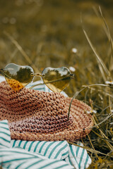 Female boho style straw hat and green heart-shaped sunglasses lie on a green grass lawn of blooming meadow. Summertime travel. Summer relax banner.