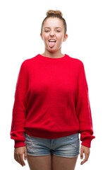 Fototapeta premium Young blonde woman wearing bun and red sweater sticking tongue out happy with funny expression. Emotion concept.