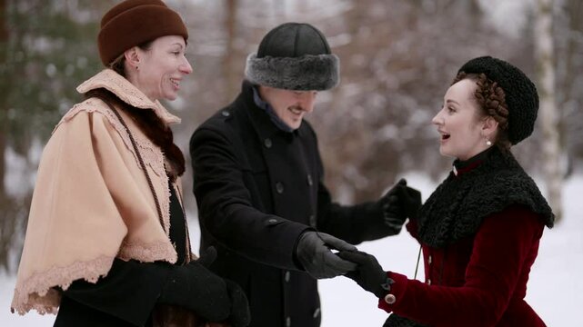 Three people in winter clothes are discussing something outdoors. The women are dressed in fur capes and hats, the man in a black coat with a fur hat.