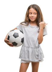 Brunette hispanic girl holding soccer football ball pointing and showing with thumb up to the side with happy face smiling