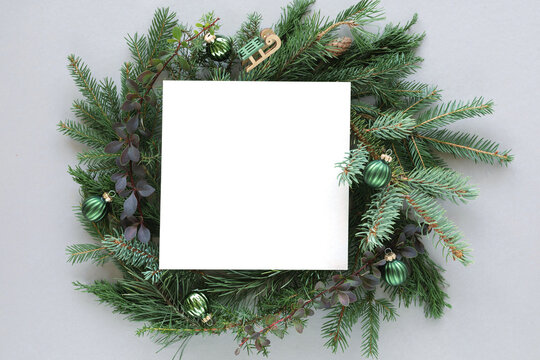 Overhead view of a blank piece of square paper in the middle of a coniferous Christmas wreath decorated with christmas baubles