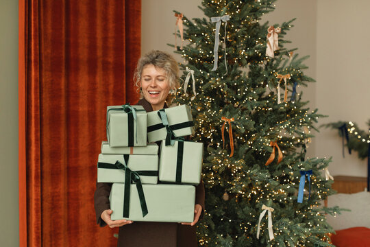 Smiling woman standing next to an illuminated Christmas tree in a living room holding a stack of wrapped Christmas gifts