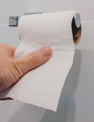 A hand pulls a sheet of toilet paper from a roll mounted on the wall in a bathroom