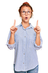 Young redhead woman wearing casual clothes and glasses amazed and surprised looking up and pointing...